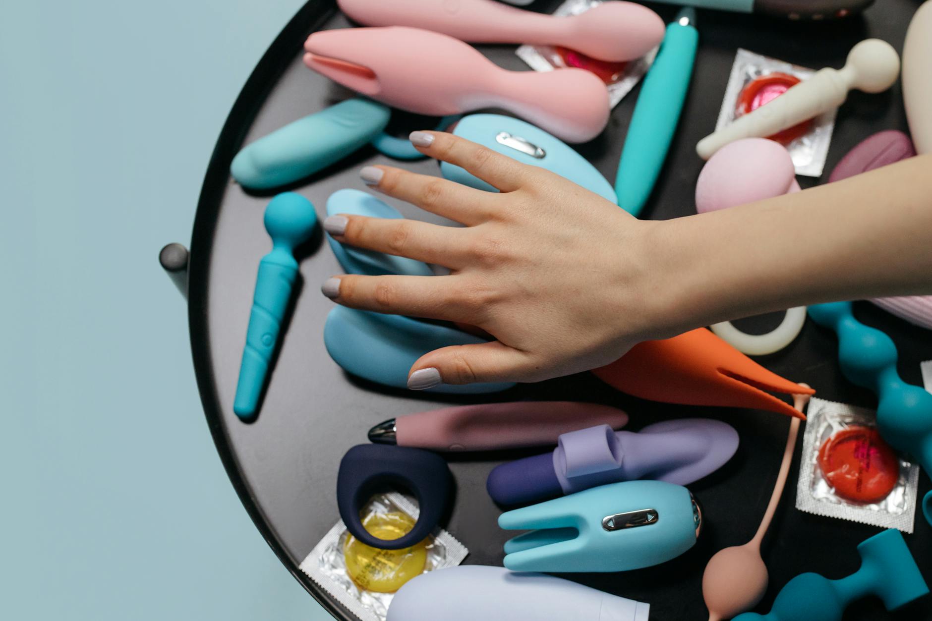A hand reaching over a variety of colorful clitoral vibrators and sex toys arranged on a surface