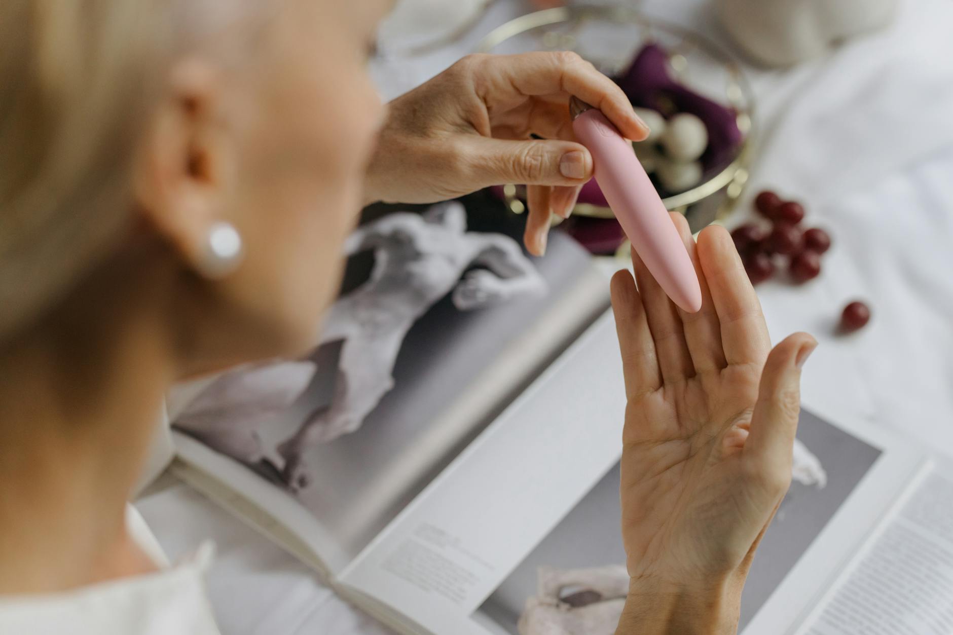 Close-up of a hand holding a pink vibrator with art books in the background, symbolizing the intersection of pleasure and personal growth.