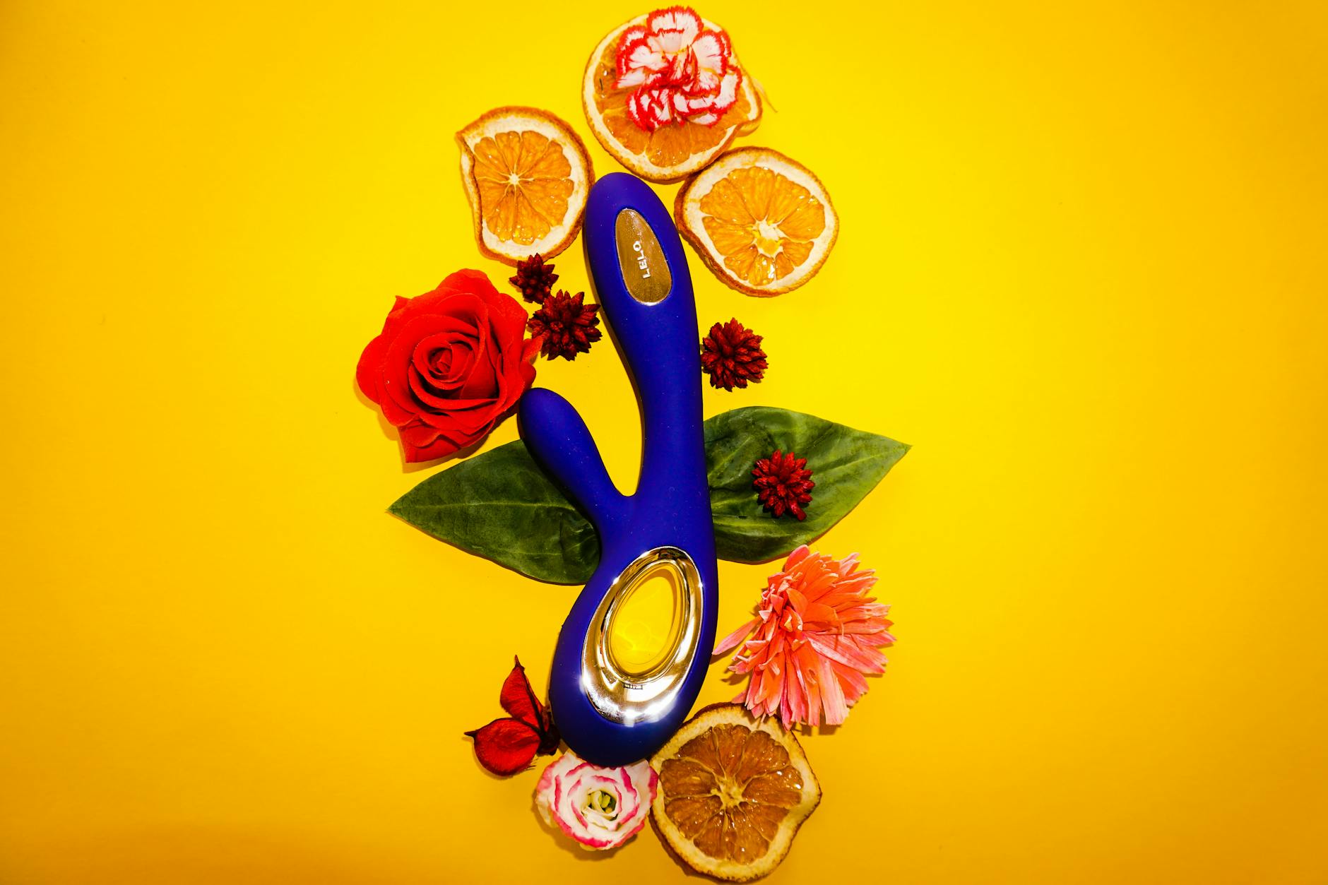 A blue clitoral vibrator surrounded by citrus and flowers on a bright yellow background